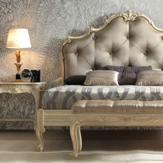 A luxurious bedroom features an ornate, tufted headboard with intricate carvings, a Small Louis Style Bench at the foot of the bed, and a side table with a decorative lamp. The bedding has a geometric design, and the wall behind displays a leafy wallpaper pattern.