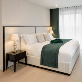 A modern bedroom design featuring a large bed with white bedding and green accents, including a green bed runner and pillows. Matching Art Deco Style Bedside Tables are on either side, each adorned with sleek lamps. Floor-to-ceiling windows with green curtains allow natural light to fill the room.