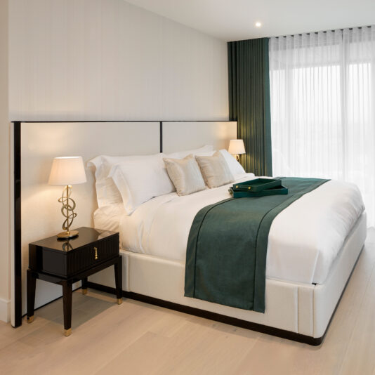 A modern bedroom design featuring a large bed with white bedding and green accents, including a green bed runner and pillows. Matching Art Deco Style Bedside Tables are on either side, each adorned with sleek lamps. Floor-to-ceiling windows with green curtains allow natural light to fill the room.