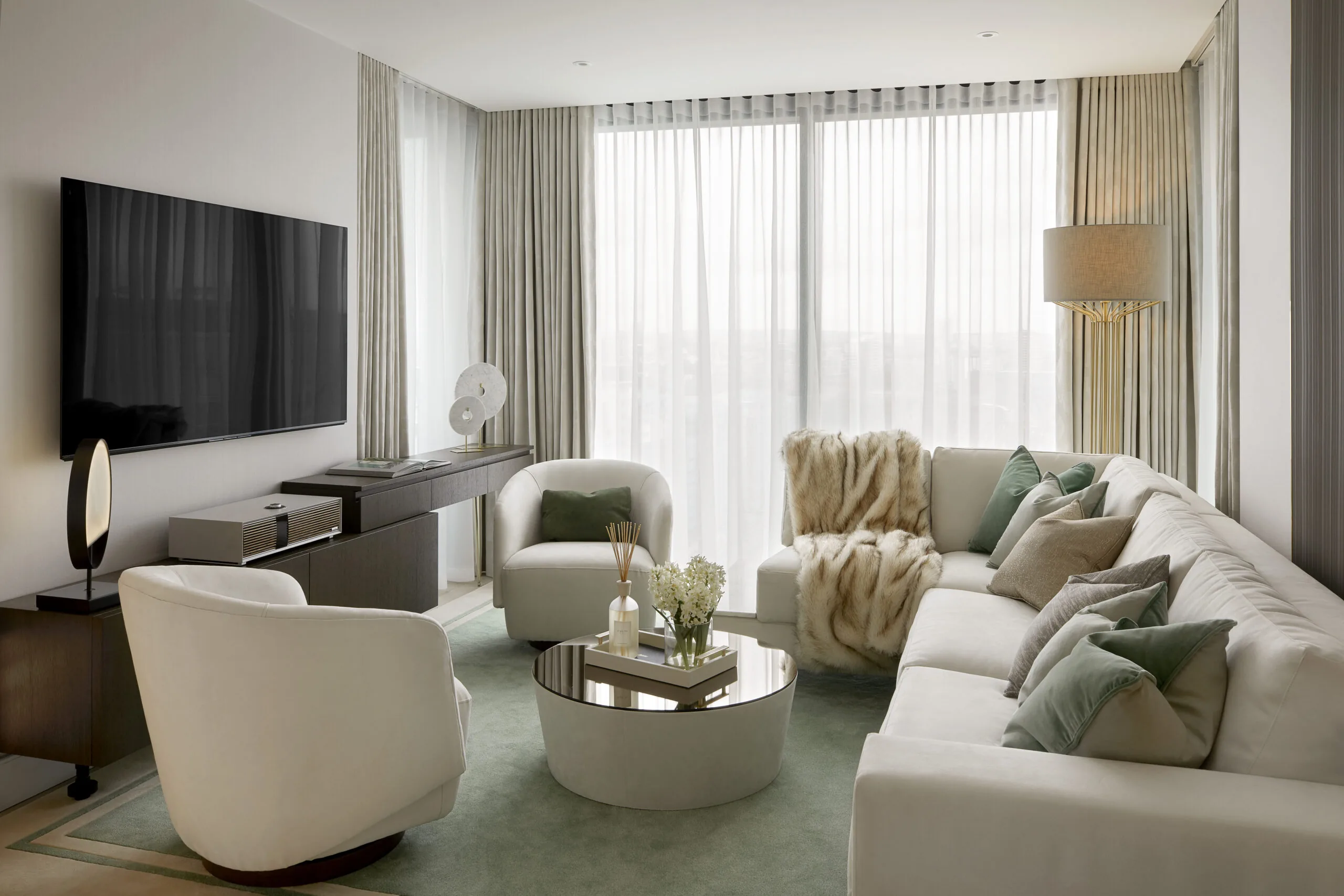 A modern living room with a large white sectional sofa, accented with green and gray pillows. A round coffee table holds decorative items. Sheer curtains cover large windows, and a flat-screen TV is mounted on a wall. Neutral tones dominate the decor.