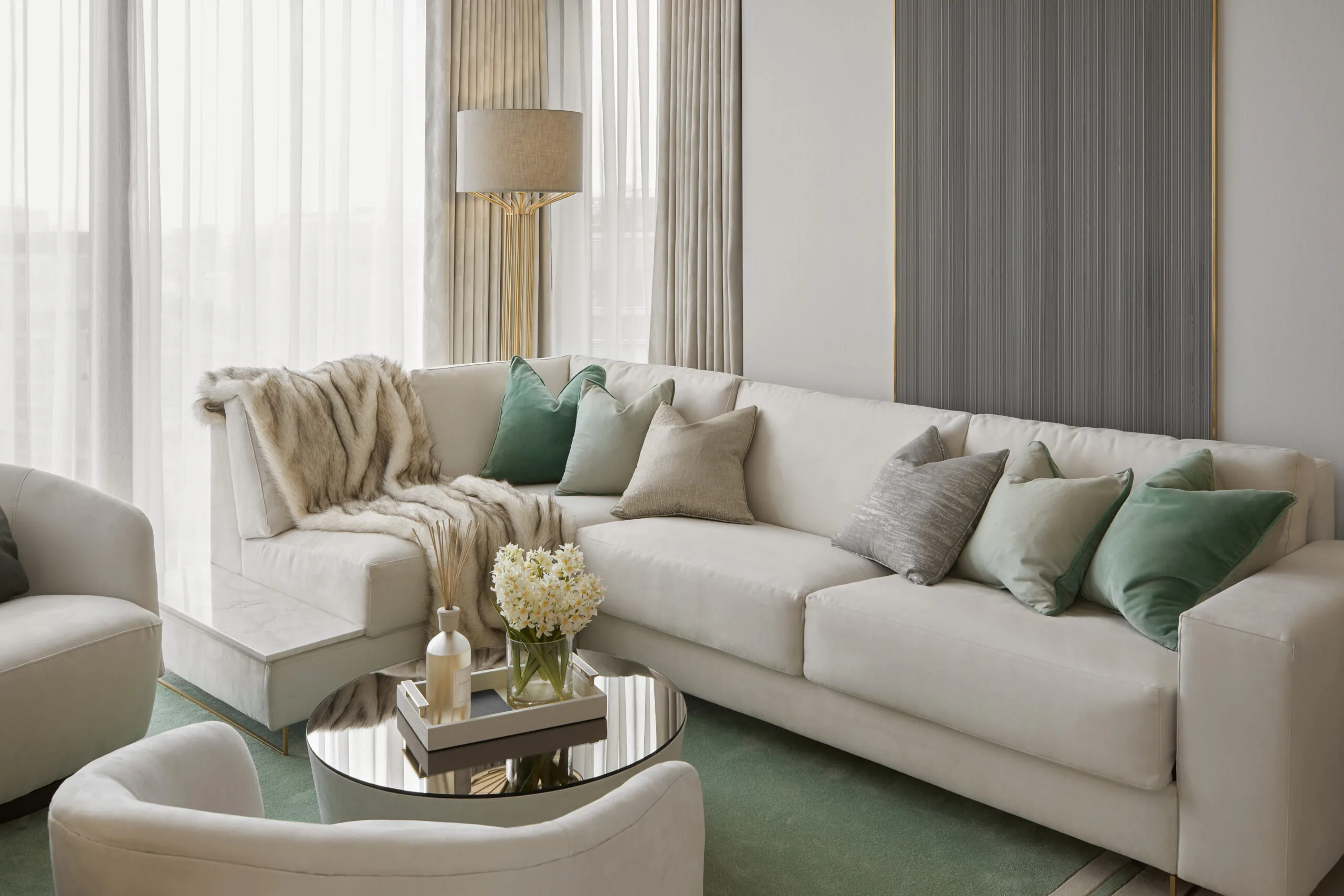 A modern living room with a large white sectional sofa adorned with gray and green cushions. A fur throw adds texture. A round glass coffee table with flowers is in front. Soft natural light filters through sheer curtains. A floor lamp stands nearby.