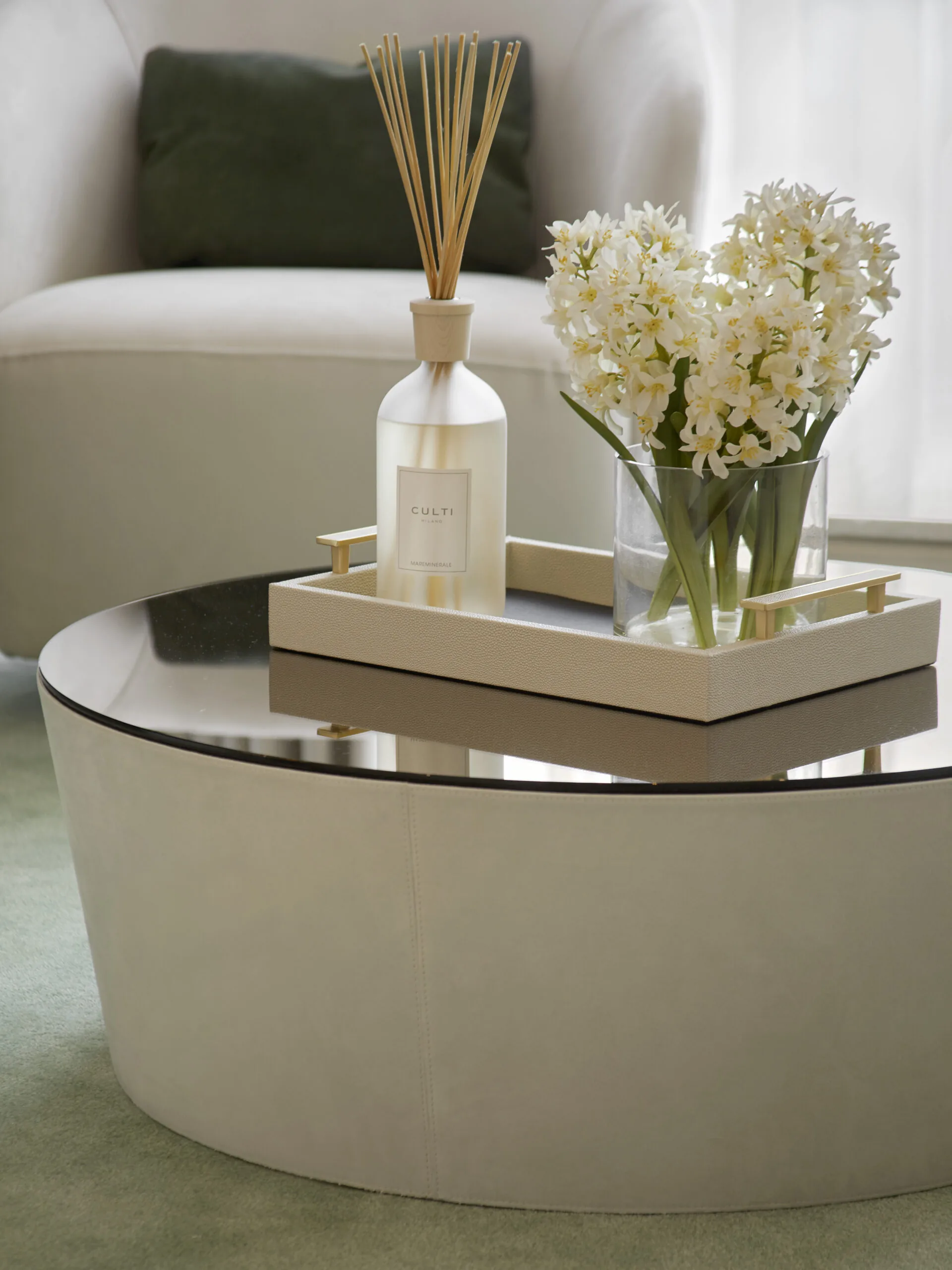 A round coffee table with a mirrored top holds a tray. On the tray are a scented diffuser with sticks and a glass vase filled with white flowers. A plush armchair with a dark green cushion is in the background. The setting is cozy and elegant.