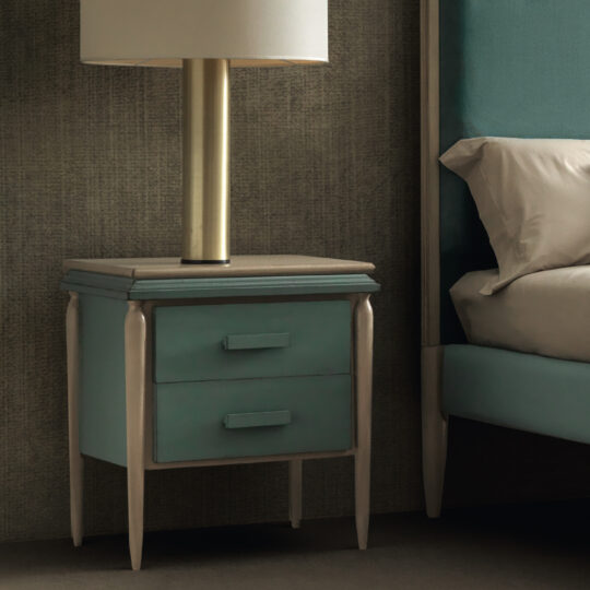 The Luxury Art Deco Style Bedside Cabinet, featuring a modern design with two drawers and a teal finish, stands next to a bed upholstered in teal. A lamp with a brass base and white shade sits on top of the cabinet. The background is adorned with textured wallpaper in a neutral tone.