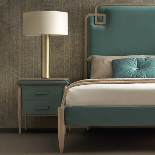 A contemporary bedroom setup featuring a bed with a teal upholstered headboard and matching bedding. To the left is the Luxury Art Deco Style Bedside Cabinet with two drawers, topped with a brass lamp featuring a white shade. The background wall has a textured, neutral tone.