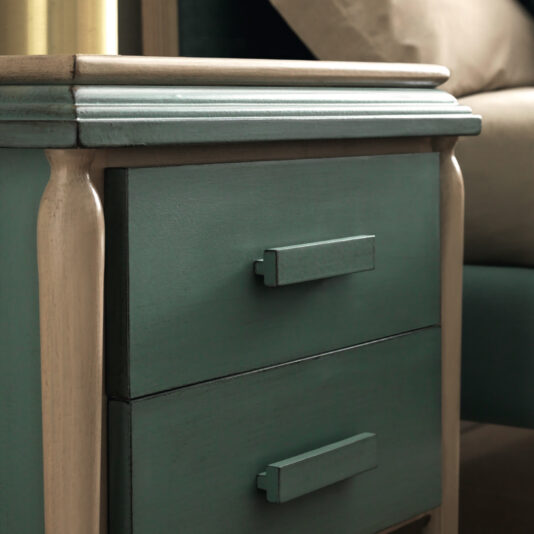 A close-up of the Luxury Art Deco Style Bedside Cabinet reveals its sophisticated design, characterized by two small drawers. Painted in elegant shades of teal and beige, it features minimalist rectangular drawer handles. Positioned next to a matching beige sofa, the top surface serves as additional storage space.