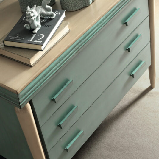 A close-up of the Luxury Art Deco Style Chest Of Drawers, a green wooden dresser with three drawers featuring horizontal handles. On top are a closed book with a dolphin figurine, another book, and a decorative ceramic item. The dresser stands on a beige carpeted floor.