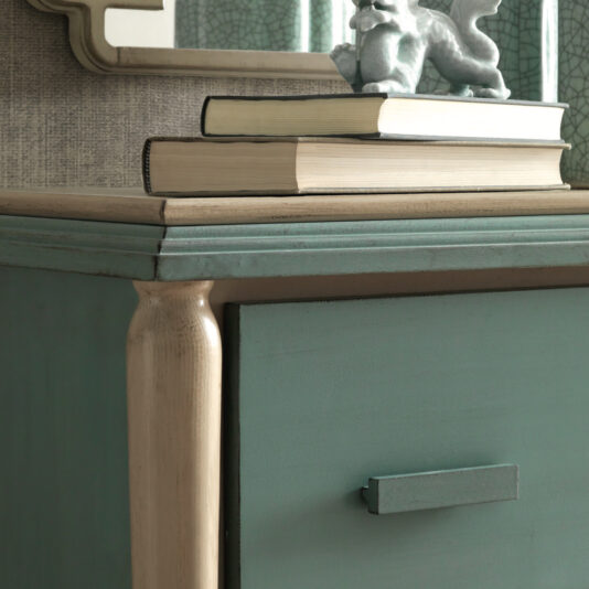 A close-up of the green Luxury Art Deco Style Chest Of Drawers with a beige handle, featuring a stack of three books and a small decorative dragon figurine on top. Behind the chest is a textured wall with an angular-framed mirror partially visible.