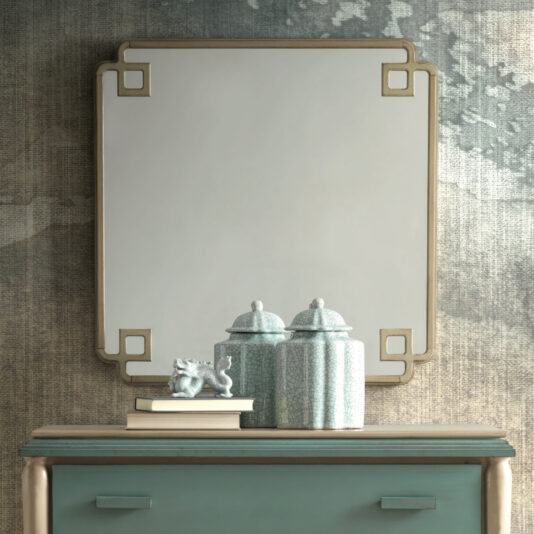 A Luxury Square Art Deco Style Mirror with elegant corners hangs on a wall; beneath it is a teal dresser adorned with two ornately designed lidded jars, a small statue, and a stack of books. The background showcases a textured, artistic wall design.