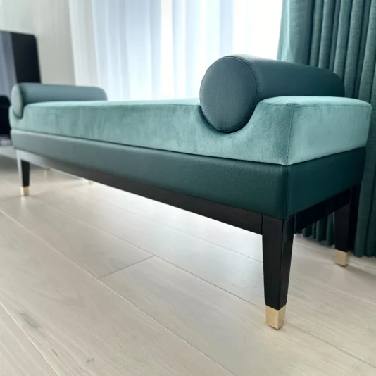 A modern teal bench with rolled armrests and sleek black legs stands on a light-colored wooden floor. A curtain hangs nearby, and natural light softly illuminates the room.