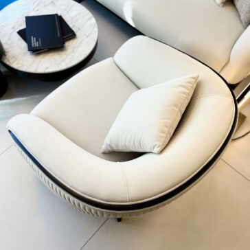 Mid Century Style Curved Armchair The Mid Century Style Curved Armchair, featuring a sleek modern design with white cushioning and a matching pillow, is elegantly placed beside a round table with a white marble top that supports two closed books. It rests beautifully on a light-colored tiled floor.
