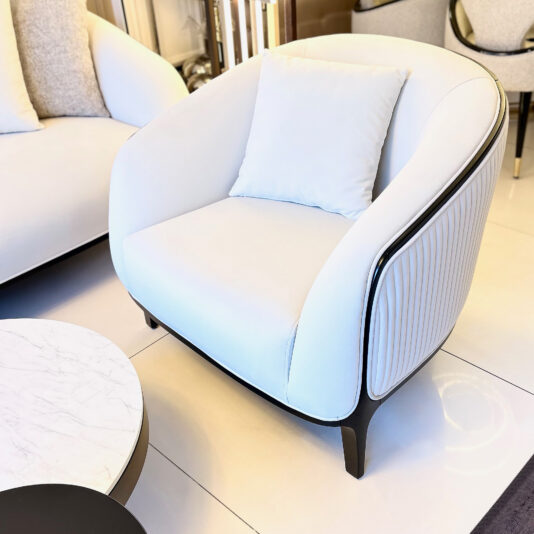Mid Century Style Curved Armchair The Mid Century Style Curved Armchair with a complementary cushion is placed beside a round marble coffee table. This chair showcases sleek black legs and ribbed side details, all set in a luminous, fashionable living room.