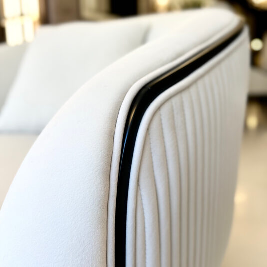 Mid Century Style Curved Armchair Close-up of the Armchair, a modern white upholstered chair with black metal accents and curved armrests. Vertical ribbed stitching highlights its sleek and elegant design against a softly blurred background.