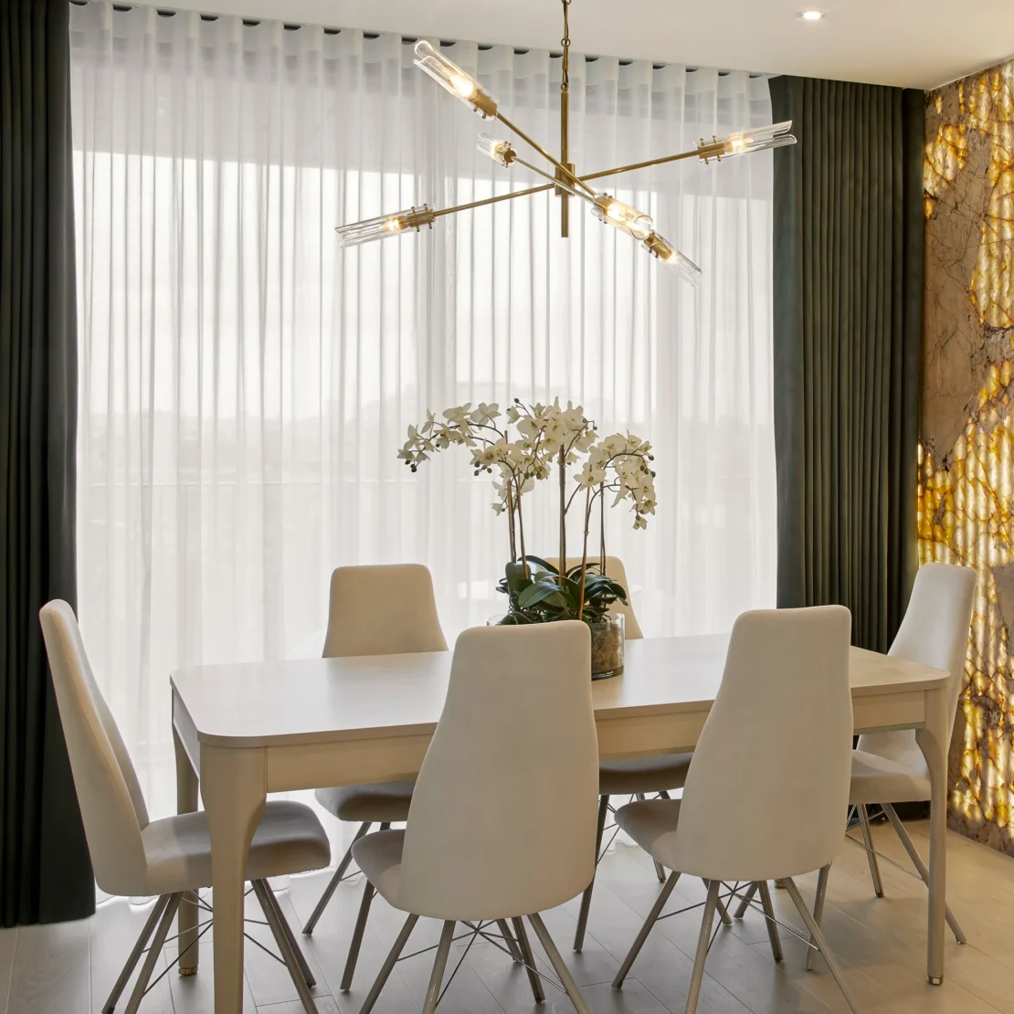 A modern dining room with a white table and six cream chairs. A gold chandelier hangs above. A vase with white orchids sits on the table. The background features large windows with sheer curtains and a stone-accented wall.