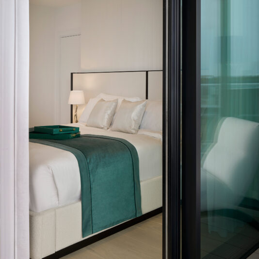 A modern hotel room featuring a Storage Bed With Curved Wide Headboard, dressed in white linens and teal accents, including a teal bed runner and matching pillows. A bedside table with a lamp is visible, and the room is partially viewed through a glass door from a balcony.