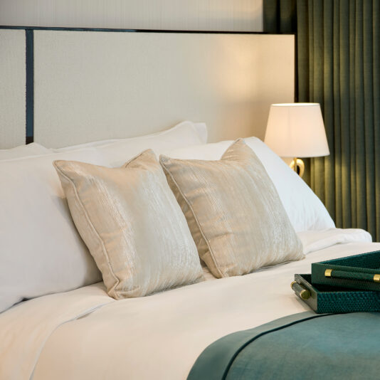 The Storage Bed With Curved Wide Headboard is neatly made with white sheets, beige accent pillows, and a teal throw blanket at the foot. A modern table lamp with a white shade is turned on, casting a warm glow, while two closed dark green books are stacked on the bed.