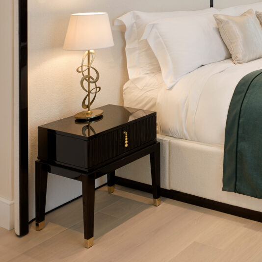 A modern bedroom nightstand with a glossy black finish, gold accents, and a small table lamp featuring a twisted gold design with a white lampshade sits beside the Storage Bed With Curved Wide Headboard, neatly made and adorned in white linens and a dark green throw blanket.