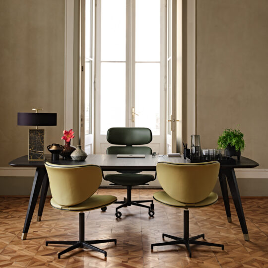 A stylish office with a Contemporary Rectangular Moka Ash Dining Table serving as the dark wooden desk, accompanied by three green chairs on a patterned wooden floor. The table features a black lamp, a potted plant, a carafe of water, a notebook, and decor items. Large windows in the background allow natural light into the room.