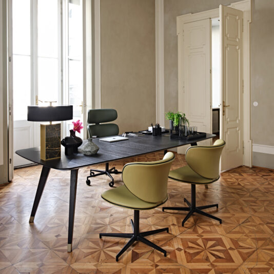 A modern office space features a sleek Contemporary Rectangular Moka Ash Dining Table with a leather chair, complemented by two green swivel chairs. Adorning the table are a lamp, plant, and decorative items. Hardwood floors and tall windows enhance the room's elegance, while open double doors provide a grand entrance to this contemporary setting.