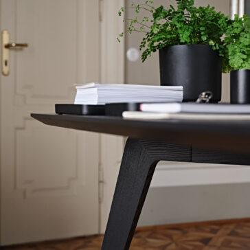 A close-up image of a contemporary black desk with a stack of neatly arranged papers, notebooks, and two potted green plants. The desk stands on a patterned wooden floor, invoking the elegance of the Contemporary Rectangular Moka Ash Dining Table. A beige door with a gold-colored handle is visible in the background.