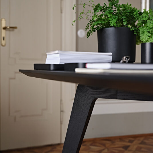 A close-up image of a contemporary black desk with a stack of neatly arranged papers, notebooks, and two potted green plants. The desk stands on a patterned wooden floor, invoking the elegance of the Contemporary Rectangular Moka Ash Dining Table. A beige door with a gold-colored handle is visible in the background.