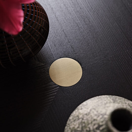 A round brass coaster lies on a dark wooden table, part of the Contemporary Rectangular Moka Ash Dining Table. A small section of a textured grey vase and a wire vase with pink flowers are visible next to the coaster.