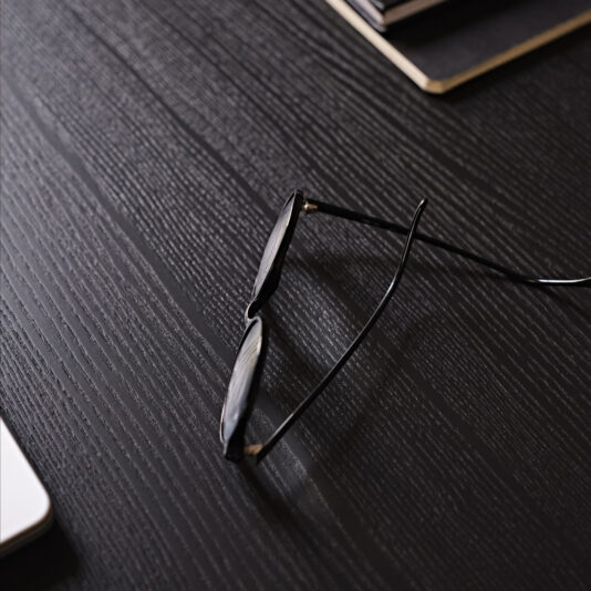 Close-up of a pair of black-rimmed glasses resting on a dark wooden desk, which complements the sleek design of a Contemporary Rectangular Moka Ash Dining Table. In the background, parts of some closed notebooks and a corner of a white laptop are visible. The scene conveys a simple, minimalist workspace aesthetic.