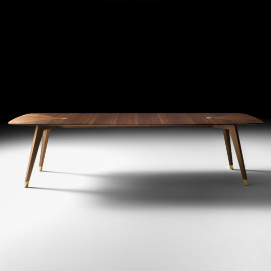The Contemporary Rectangular Walnut Dining Table is showcased in a brightly lit room with a dark background. This modern piece features tapered legs, each accented with metallic caps on the tips. The tabletop boasts a smooth finish, and its slightly rounded edges contribute to its sleek and contemporary design.