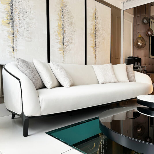 In a contemporary living room, the Mid Century Style Curved Sofa takes center stage with its sleek white design, enhanced by light gray cushions. The wall behind is adorned with abstract art pieces featuring vertical gold and gray accents, while a glass coffee table brings the decor together.
