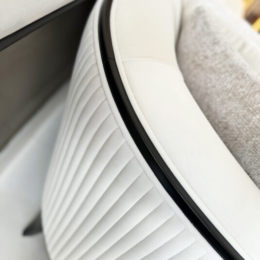 Close-up of a modern, white upholstered chair featuring vertical stitching on the backrest. Its sleek black frame outlines the chair while a textured, light gray cushion adds to its appeal. The minimalist design exudes elegance reminiscent of the timeless allure found in the Mid Century Style Curved Sofa.