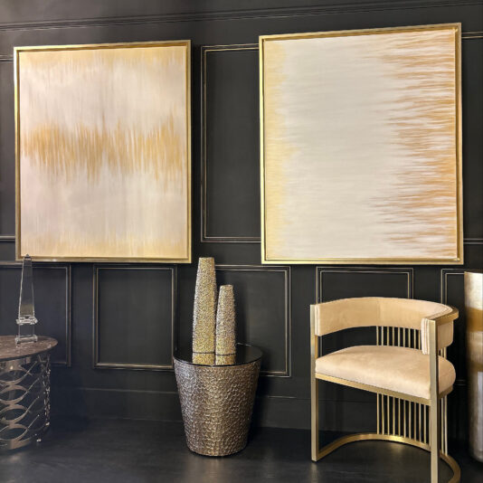 A chic interior featuring two copies of the Original Gold Leaf Abstract Painting on a black paneled wall. Below, a metallic textured planter with tall decorative vases sits next to a beige upholstered chair with a gold frame. An elegant side table stands on the left.