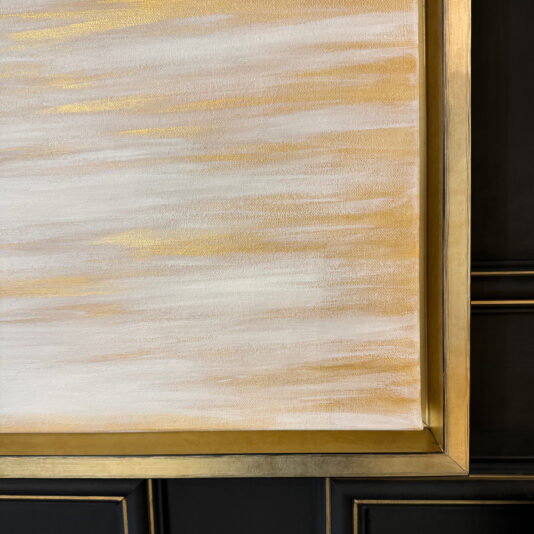 Pair Of Original Abstract Paintings A close-up shot of the Original Gold Leaf Abstract Painting (Copy) with a gold metallic frame. The artwork showcases horizontal brush strokes in shades of white, cream, and gold, creating a soothing, textured appearance. The background features part of a dark green wall with paneling.