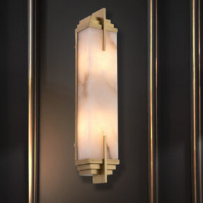An elegant wall sconce with an elongated, rectangular shape, featuring a frosted glass center emitting a warm glow. It's framed by gold-toned geometric accents, mounted on a dark wall with subtle vertical lines.