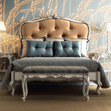 Carved Louis Style Tufted Bed The bedroom exudes luxury with a Classic Louis Style Button Upholstered Bed, showcasing a tufted headboard adorned with blue pillows. A coordinating bench rests at the foot, complemented by wheat-patterned wallpaper and two lamps atop intricately designed side tables, completing the elegant decor.
