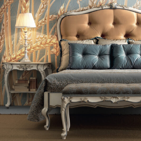 Carved Louis Style Tufted Bed An elegant bedroom features the Carved Louis Style Tufted Bed, beautifully adorned with blue and gold pillows. A vintage-style nightstand next to it supports a lit lamp, while the wallpaper displays a close-up wheat design, adding a rustic touch.