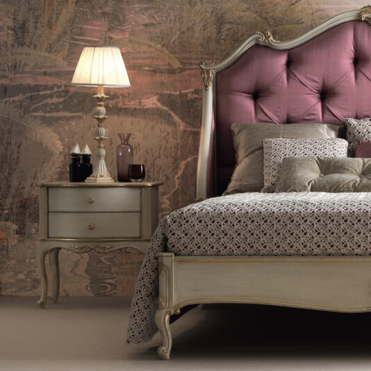 A luxurious bedroom featuring an elegant, classic-style bed with a tufted purple headboard and patterned bedding. Beside it is the Elegant Classic Style Bedside Drawers, a vintage-style nightstand with two drawers holding a tall, exquisite lamp and decorative items. The wall has a textured, artistic design.