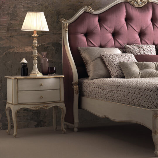 An elegant bedroom scene with a classic style features a tall, ornate headboard adorned with tufted maroon upholstery. The bed itself is festooned with multiple pillows and a patterned comforter. Beside it, the Elegant Classic Style Bedside Drawers hold a decorative lamp and various items.