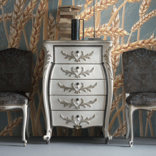 A sophisticated silver dresser adorned with intricate leaf designs on its drawers, similar to the Classic Style 5 Drawer Tallboy, is positioned between two dark upholstered chairs. On top, it holds stacked books and a vase. The setting is enhanced by a wheat-themed wallpaper in the background.