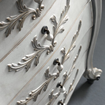 Classic Style 5 Drawer Tallboy A close-up of the Classic Style 5 Drawer Tallboy showcases its vintage charm, featuring ornate floral carvings and elegant metallic handles. Painted in a soft, distressed white, the wood finish accentuates the intricate details. The dark floor provides a striking contrast to this light-toned piece of furniture.