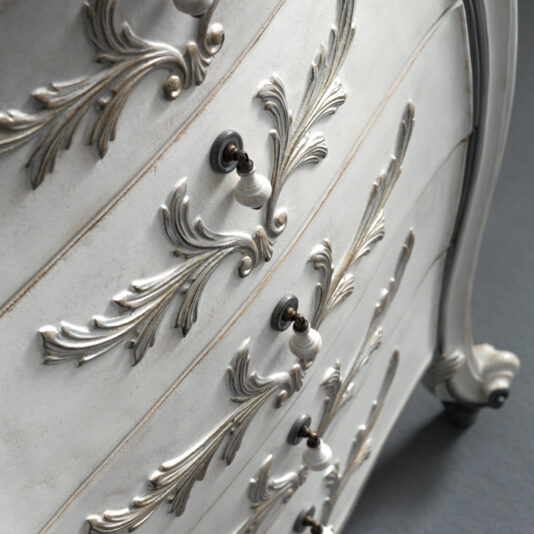 A close-up of the Classic Style 5 Drawer Tallboy showcases its vintage charm, featuring ornate floral carvings and elegant metallic handles. Painted in a soft, distressed white, the wood finish accentuates the intricate details. The dark floor provides a striking contrast to this light-toned piece of furniture.