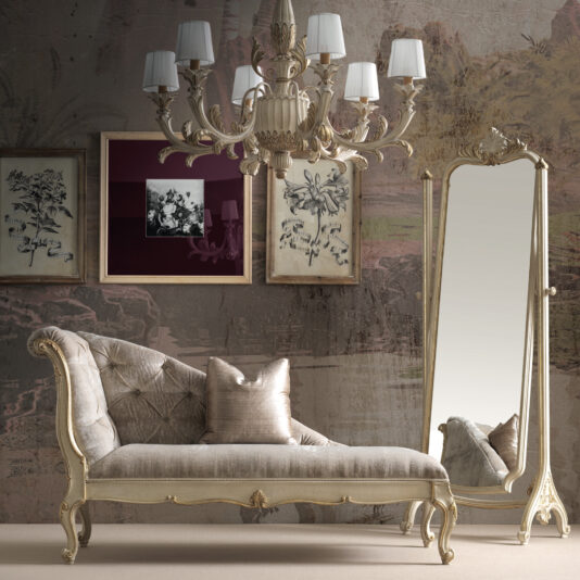 A luxurious room features a vintage chaise lounge with tufted cushions, an ornate floor mirror, and botanical wall art. The Classic Designer Ornate Italian 6 Arm Chandelier with lampshades hangs elegantly above against a textured, earthy wall backdrop.