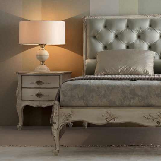 A bedroom scene featuring a stylish, tufted headboard attached to a light-colored bed. The bed has a matching comforter and pillows. Beside it, there's an Ornate Classic Style Bedside Table with two drawers, topped with a ceramic lamp emitting a warm light. The walls have a textured, gradient pattern.