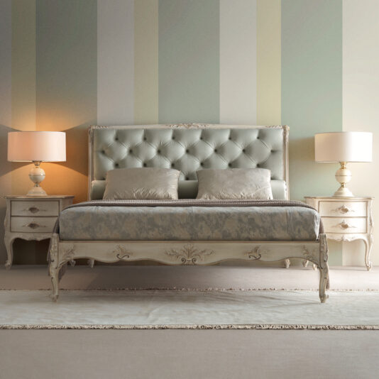 Elegant Classic Style Bedside Drawers A stylish bedroom features a king-size bed with a tufted headboard, adorned with decorative pillows and a luxurious bedspread. Flanking the bed are two Ornate Classic Style Beside Tables, each topped with a lit table lamp. The room's walls display green and beige striped wallpaper.