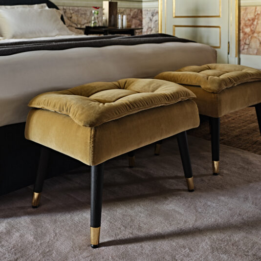 Two plush, mustard-colored ottomans with black legs sit on a light gray carpet beside a luxurious bed. The room features elegant decor with marble accents and a hint of a neatly made bed in the background.