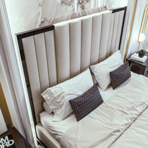 A luxurious bedroom features a bed with a tall, padded beige headboard. The bed is neatly made with white linens and patterned accent pillows. A nightstand with a lit lamp and decor is visible, with a hint of marble wall detail behind.