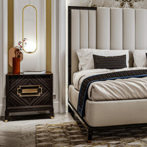 A stylish bedroom scene features a plush beige bed with patterned pillows and a blue decorative throw. A dark wooden nightstand with a geometric design holds a vase and a modern wall lamp. A patterned rug adds warmth to the floor.