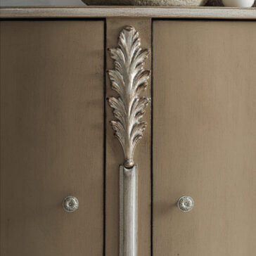 A close-up view of the High End Rococo Inspired Sideboard showcases its luxurious beige finish, featuring ornate metallic leaf-shaped handles and two circular knobs. The intricate design is accentuated by soft lighting that highlights the texture and craftsmanship of this exquisite cabinet.
