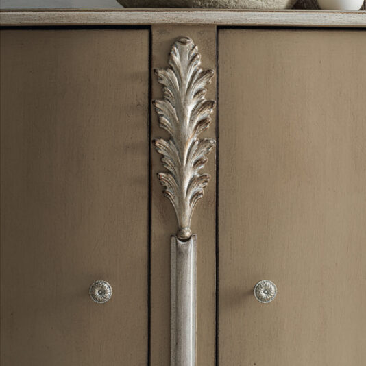 High End Rococo Inspired Sideboard A close-up view of the High End Rococo Inspired Sideboard showcases its luxurious beige finish, featuring ornate metallic leaf-shaped handles and two circular knobs. The intricate design is accentuated by soft lighting that highlights the texture and craftsmanship of this exquisite cabinet.
