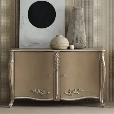 A High End Rococo Inspired Sideboard, with its vintage gold-toned finish and intricate floral carvings, stands against a textured wall. On top, two vases and a round black artwork complement its elegant design, creating an antique appearance.