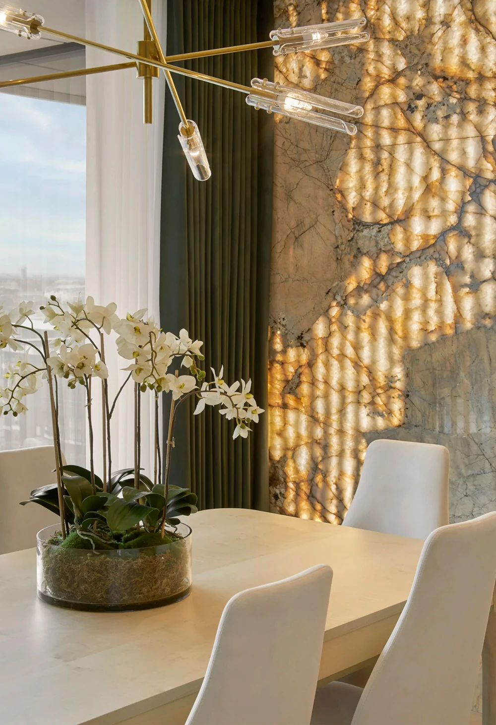 A modern dining area in this Apartment Battersea features a light wood table with white chairs. A glass pot of white orchids serves as the centerpiece. The wall showcases a striking backlit marble design, while a contemporary chandelier with exposed bulbs hangs above the table.