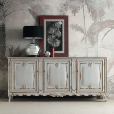 A Large Rococo Inspired Sideboard with intricate details is positioned against a wall adorned with tropical wallpaper. Atop the sideboard, there is a sleek modern silver lamp, a framed black-and-white artwork set against a burgundy mat, and a small vase. The entire arrangement seamlessly combines classic and contemporary elements.